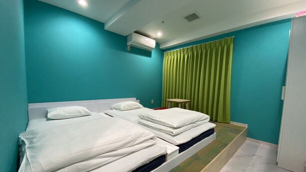 Habitación de hotel con dos camas, paredes color turquesa, una cortina verde y una mesita.