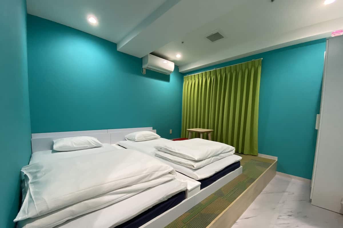 Quarto de hotel com duas camas, paredes verde-água, cortina verde e uma pequena mesa.