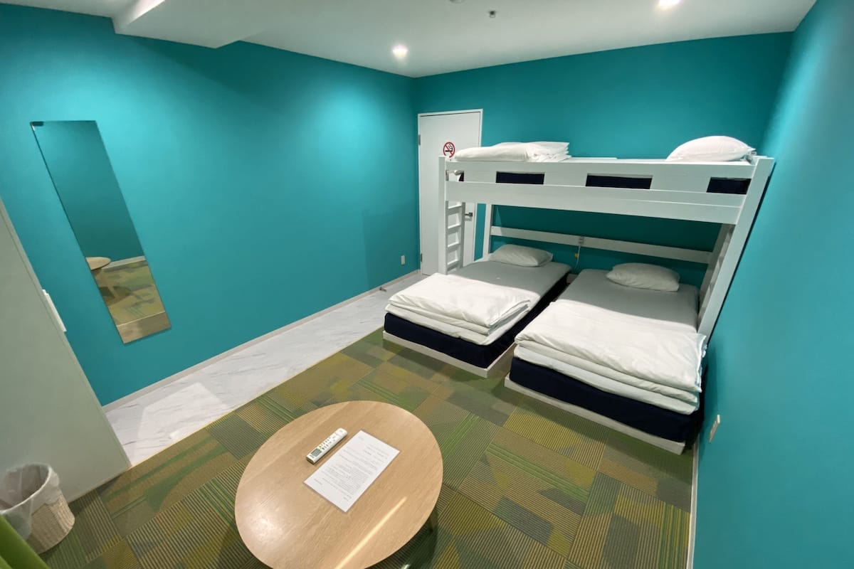 Quarto com duas beliches, uma mesinha redonda com um controle remoto e um papel, e uma porta com a placa 'Proibido Entrar'.