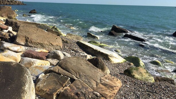 A rocky shoreline with large boulders and pebbles, leading to calm sea waters.