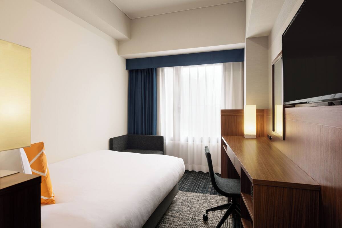 Habitación de hotel con cama, escritorio, silla, televisor y ventana con cortinas.