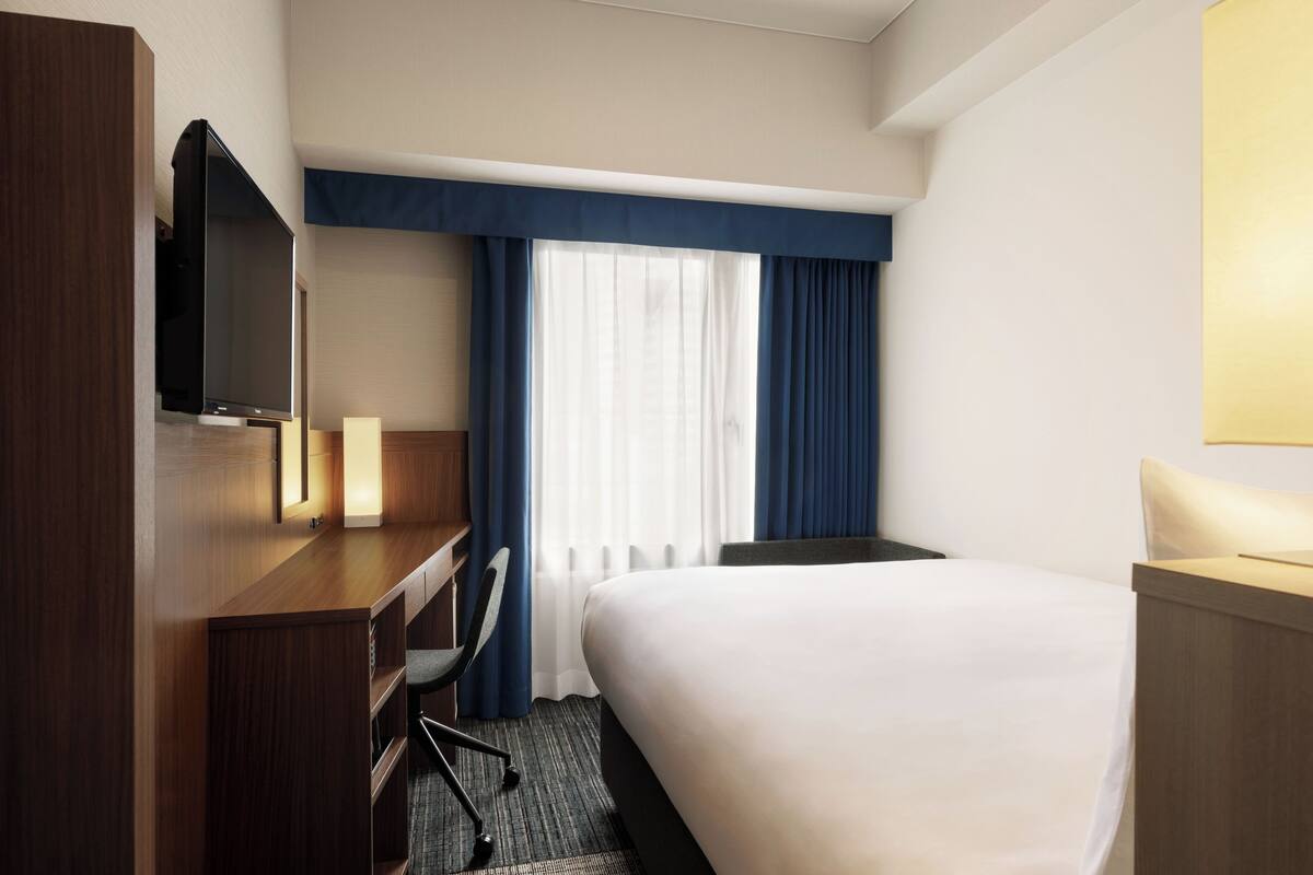 Habitación de hotel con cama, escritorio con silla, televisor y ventana con cortinas.