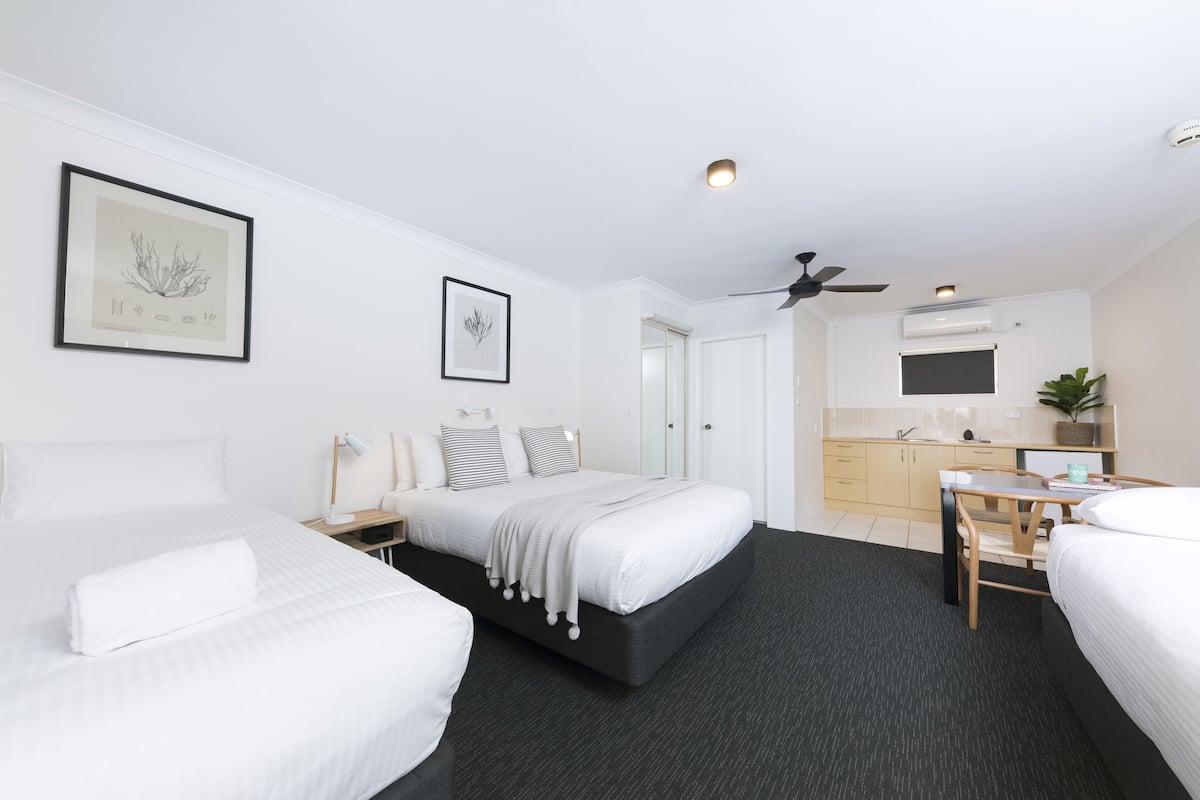 Habitación de hotel con dos camas, un escritorio, una silla, ventilador de techo y una kitchenette.