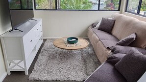 Uma sala de estar moderna com sofá em formato de L, televisão de tela plana sobre um armário branco, mesa de centro redonda e grandes janelas com vista para o verde lá fora.