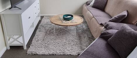 Un salon moderne avec un canapé d’angle, un téléviseur à écran plat posé sur un meuble blanc, une table basse ronde et de grandes fenêtres donnant sur un paysage verdoyant.