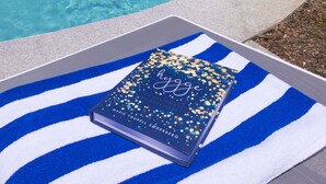 Uma área de piscina com espreguiçadeiras e guarda-sóis, um livro sobre uma toalha, e uma placa de hotel ao fundo.