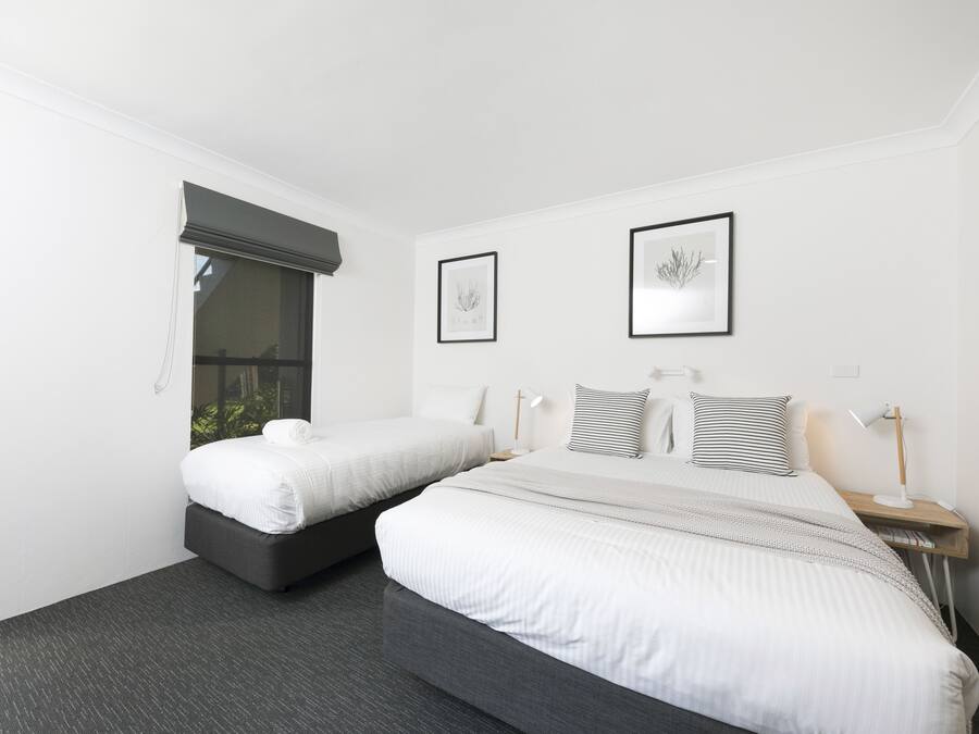 Ein Hotelzimmer mit zwei Betten, einem großen Fenster mit Verdunkelungsvorhang, zwei gerahmten Kunstwerken an der Wand und einem Nachttisch mit Lampe.