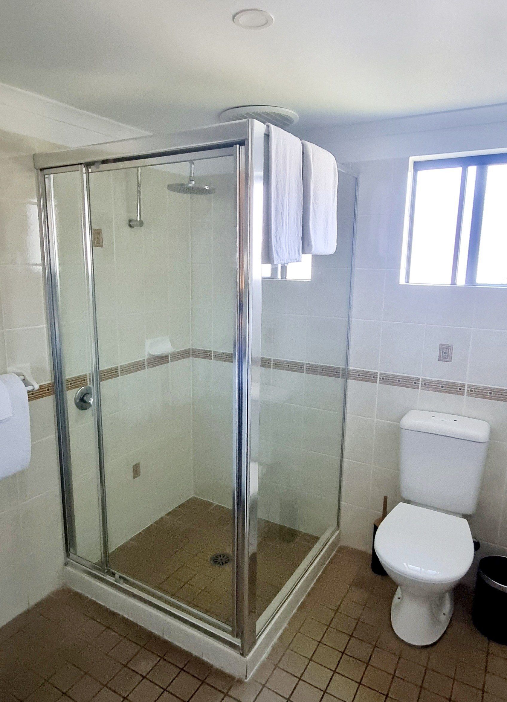 Et badeværelse med et glasafskærmet brusebad, toilet og et vindue der giver naturligt lys.