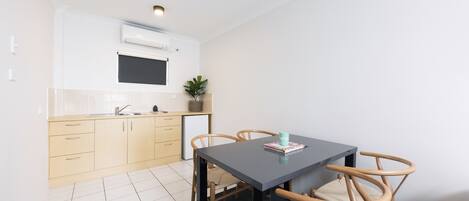 A modern kitchen with white cabinets, a black countertop, and a small refrigerator. There is a dining table with four chairs and a plant on the counter.