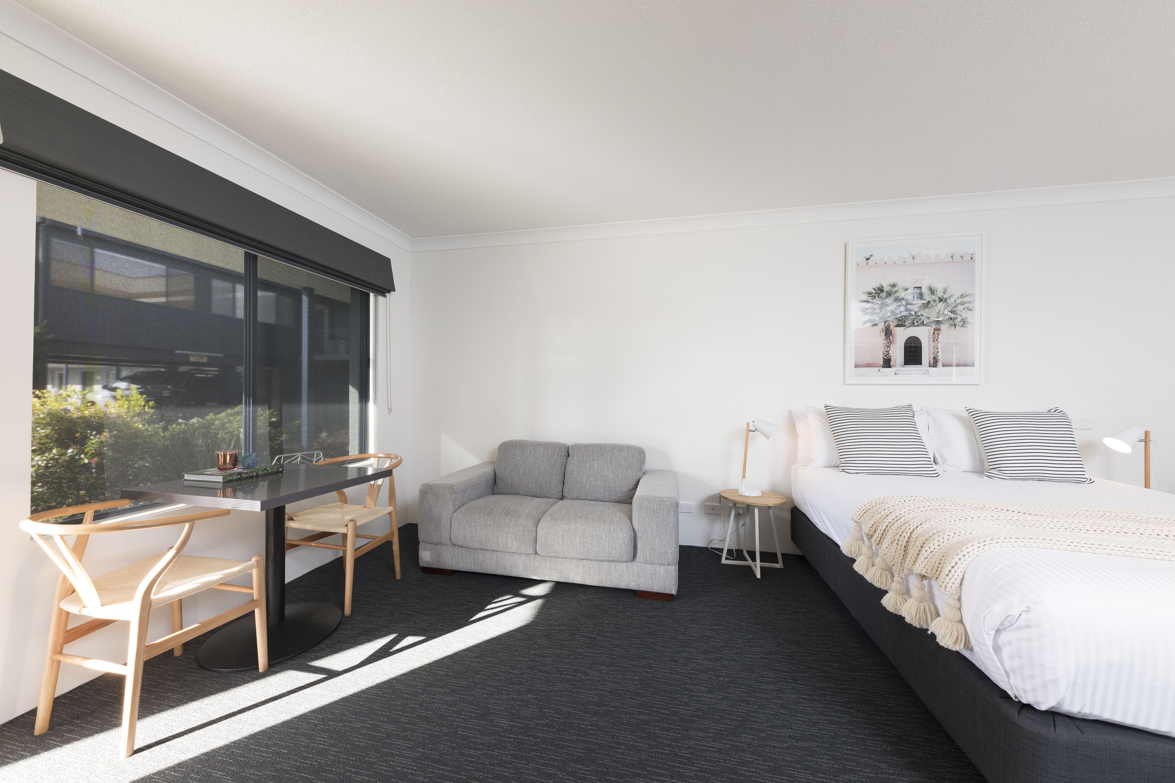 A modern hotel room with a large window, a gray sofa, a small table, a bed with white bedding, and a framed picture on the wall.