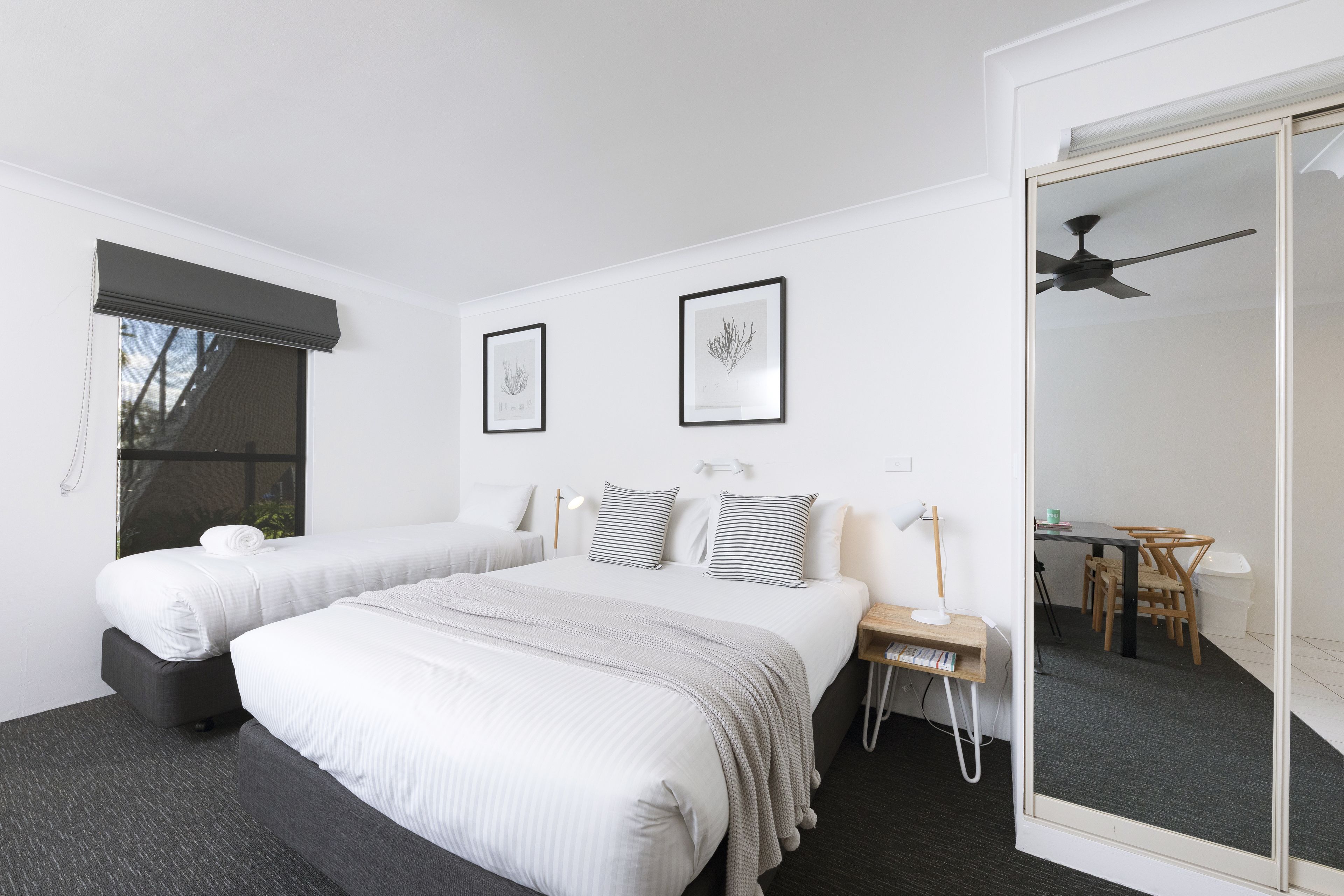 A modern hotel room with two beds, a desk, a chair, a mirror, and a ceiling fan.