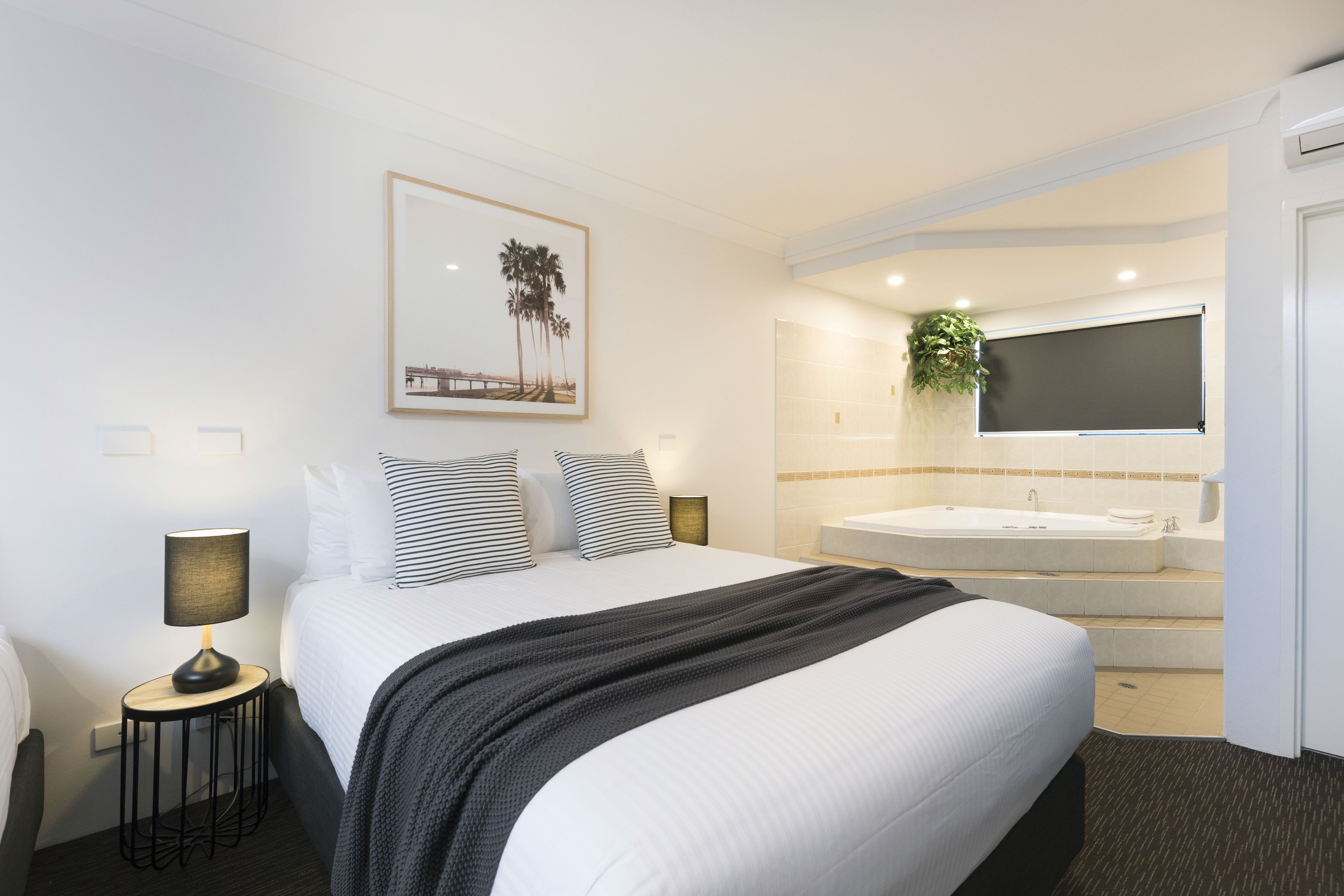 Habitación de hotel con una cama grande, una mesita de noche con lámpara, un cuadro en la pared, un ventanal amplio y un baño con bañera y lavamanos.
