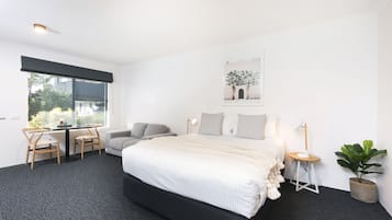 A modern bedroom with a large bed, a desk with two chairs, a small table with a lamp, and a potted plant.