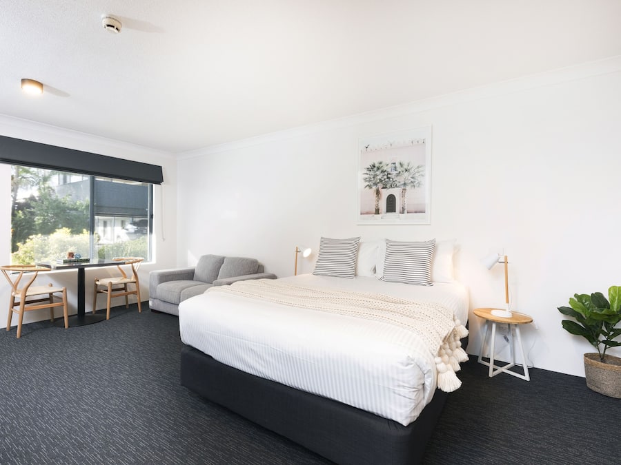 A modern bedroom with a large bed, a desk with two chairs, a small table with a lamp, and a potted plant.
