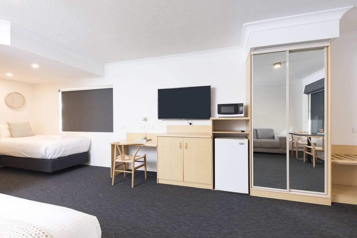 Una habitación de hotel moderna con cama, escritorio, armario, televisor, microondas y espejo.