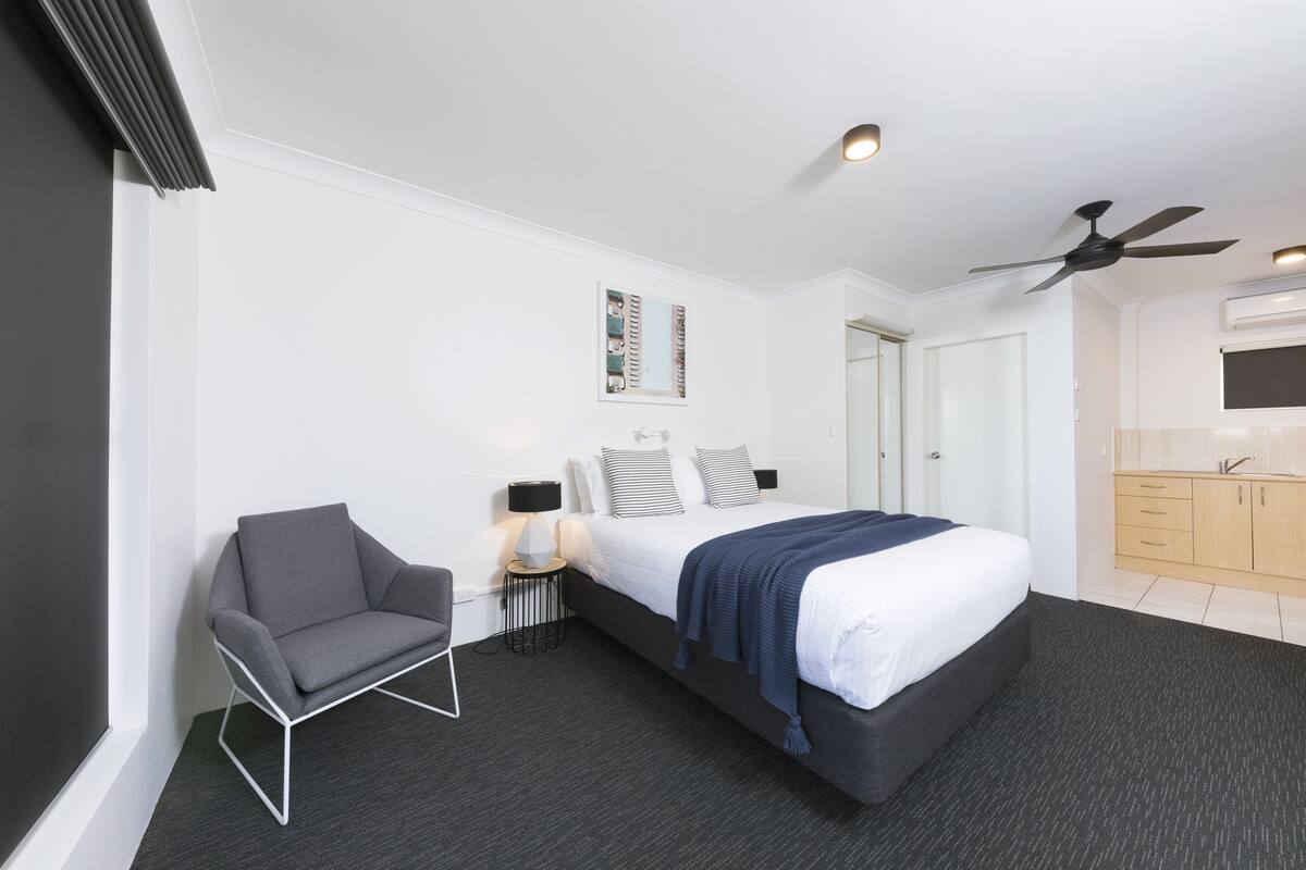 Una habitación de hotel moderna con una cama grande, un sillón gris, una mesita auxiliar pequeña y un ventilador de techo.