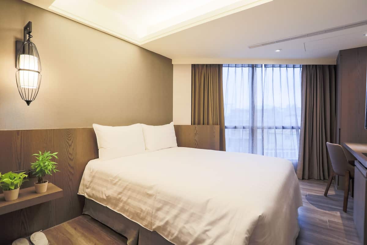 A hotel room with a large bed, a bedside table with a lamp, a small plant, and a view of the city through the window.