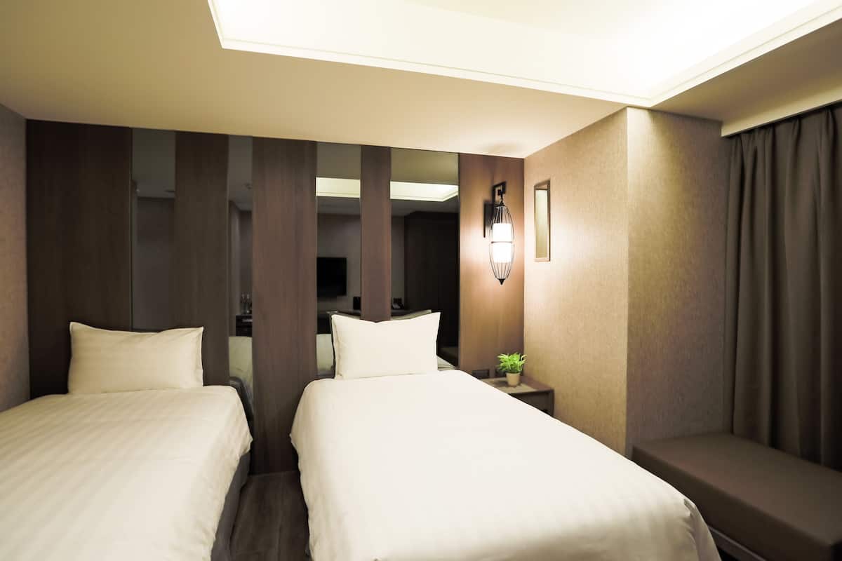 A hotel room with two beds, a bedside table with a lamp, and a large mirror.