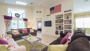 Ein Wohnzimmer mit einer Couch, Sesseln, einem Couchtisch und einem Fernseher. Ein Bücherregal mit Dekorationselementen und ein Fenster mit Vorhängen.