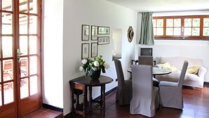 Ein Essbereich mit Tisch, Stühlen und einer Couch. An der Wand hängen Bilder und es gibt ein Fenster mit Vorhängen.