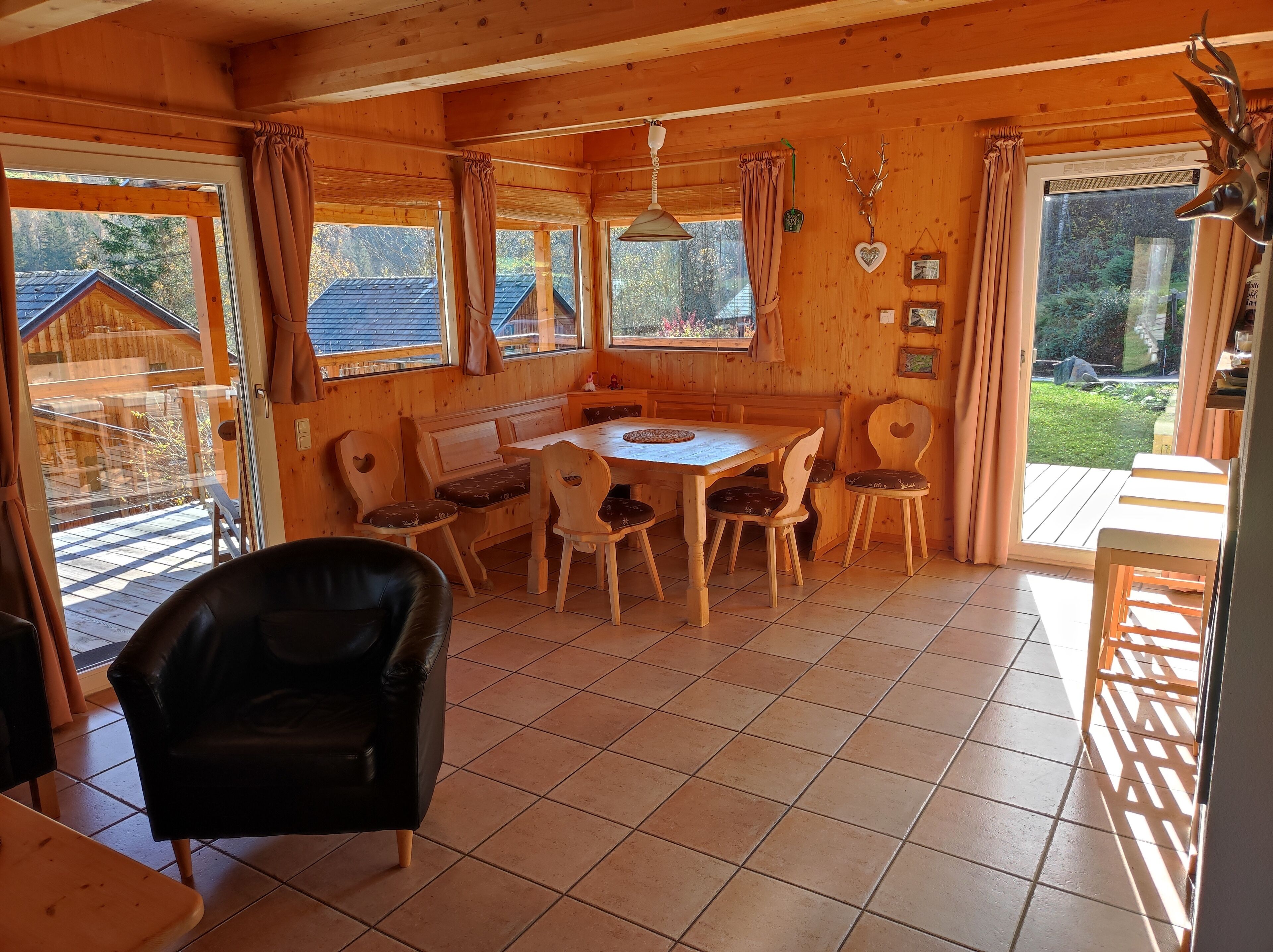 A wooden cabin interior with a dining area, wooden chairs, a bench, and a view of a wooden house outside.