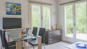 Ett modernt matområde med ett glasbord, svarta stolar, en tv och utsikt över grönskan genom stora skjutdörrar.