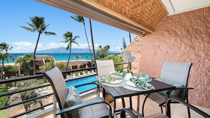 Un balcón con una mesa lista para comer, con vista a una piscina y una playa con palmeras y montañas a lo lejos.