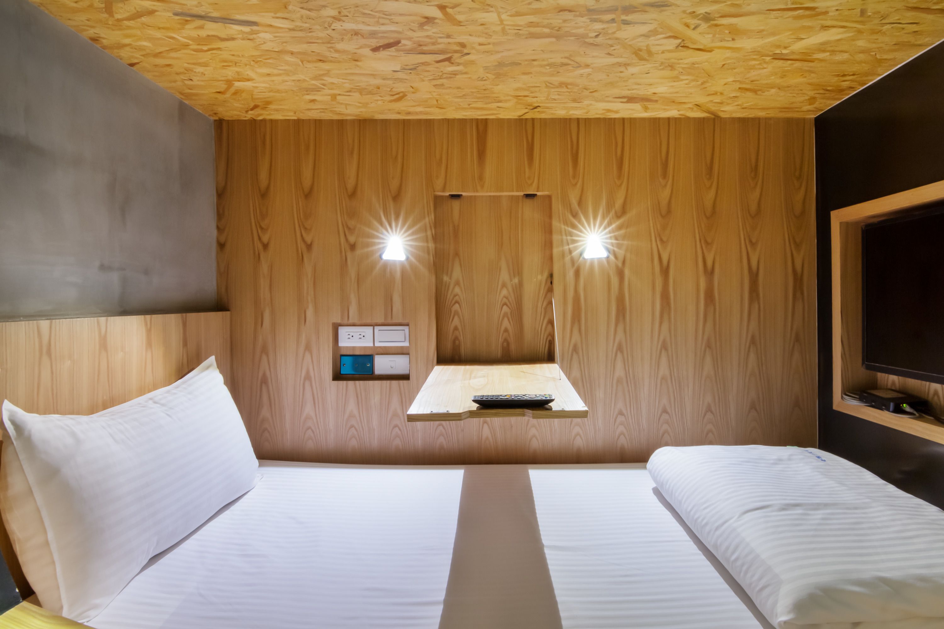 A room with two beds, wooden panel walls, a small table with a remote control, and a wall-mounted TV.