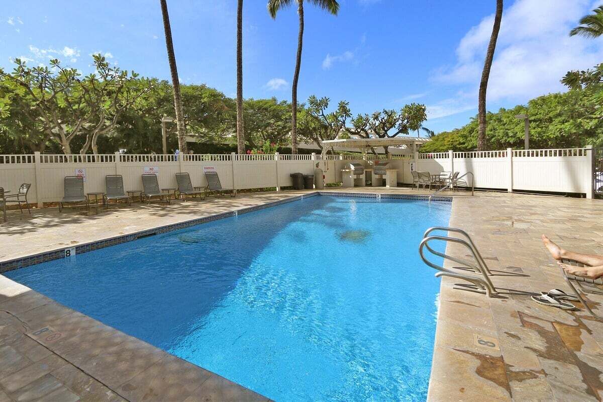 A swimming pool with lounge chairs and a covered area, surrounded by palm trees and a white fence.