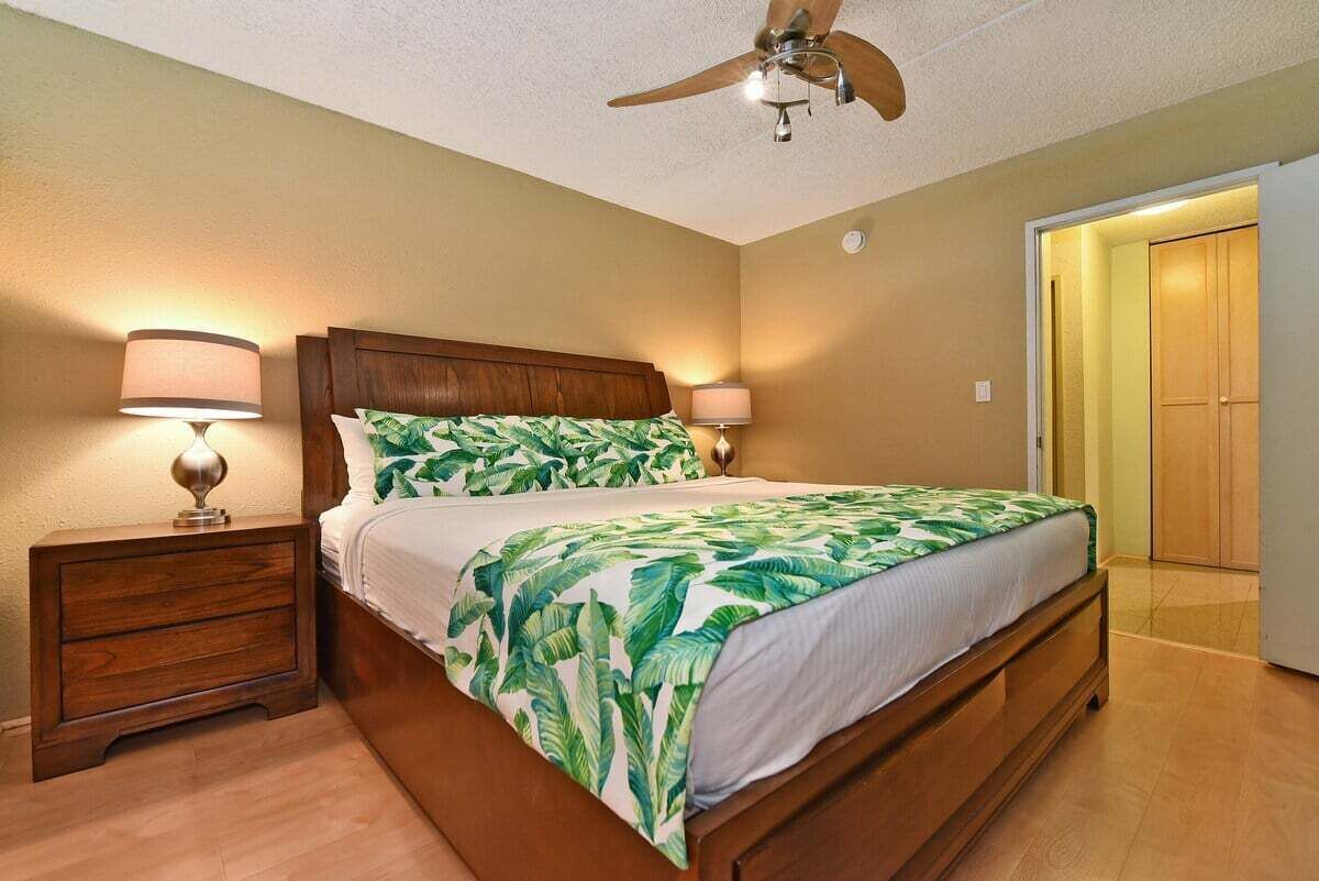 Un dormitorio con cama, mesitas de noche, lámparas, ventilador de techo y una puerta que da a otra habitación.