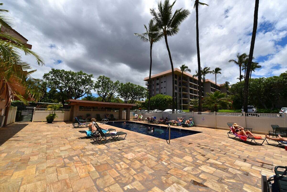Zona de piscina con sillas de descanso, palmeras y un edificio al fondo.