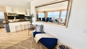 A modern living room with a large mirror, a blue sofa, and a view of the kitchen and dining area. - 5 BR Large Beach House, Beachfront with Fire Ring,  Walk to Restaurants (Oceanside)