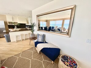 A modern living room with a large mirror, a blue sofa, and a view of the kitchen and dining area. - 5 BR Large Beach House, Beachfront with Fire Ring,  Walk to Restaurants (Oceanside)