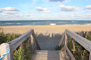 Un sentier en bois mène à une plage de sable, avec l’océan en arrière-plan.