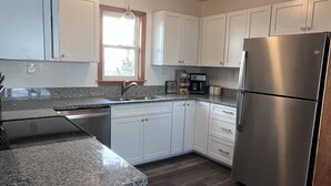 A modern kitchen with white cabinets, a stainless steel refrigerator, granite countertops, and a window providing natural light. - OBX, NC, Pristine Ocean Views without oceanfront price! (Kitty Hawk)