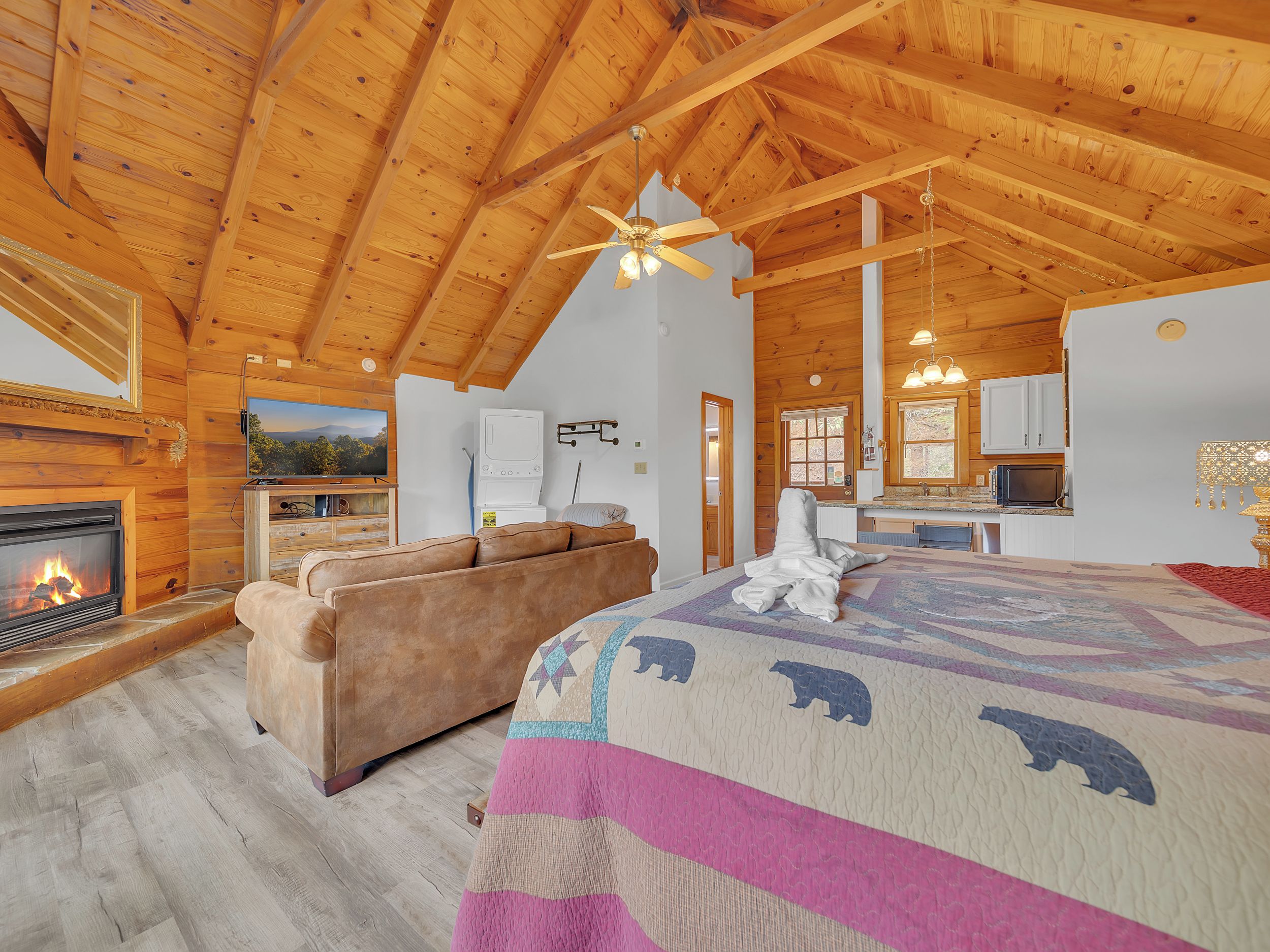 A log cabin interior with a fireplace, sofa, bed with a bear quilt, wooden ceiling, and a kitchen area.