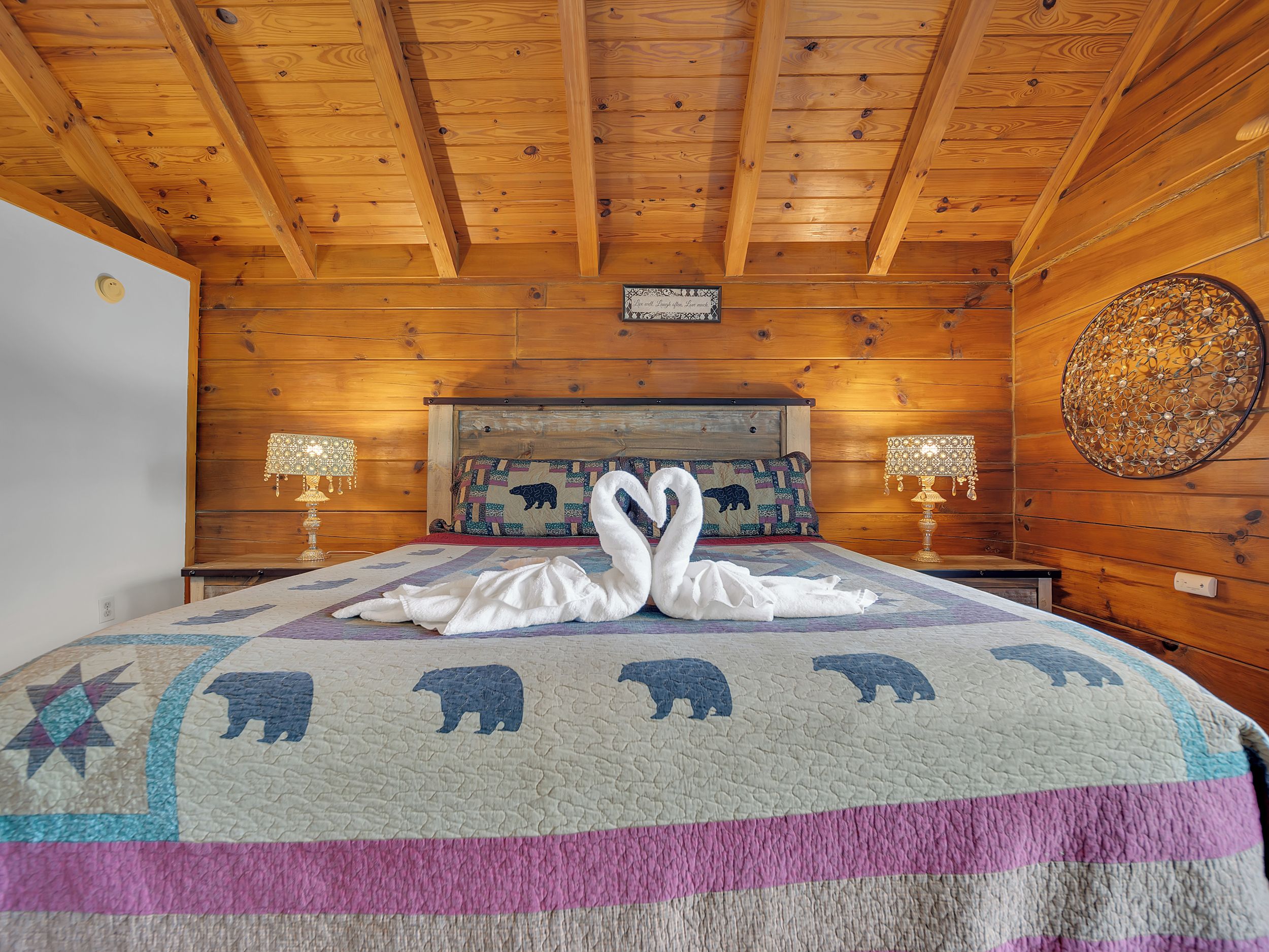 A wooden cabin room with a bed, two bedside lamps, a decorative wall piece, and a sign above the bed.