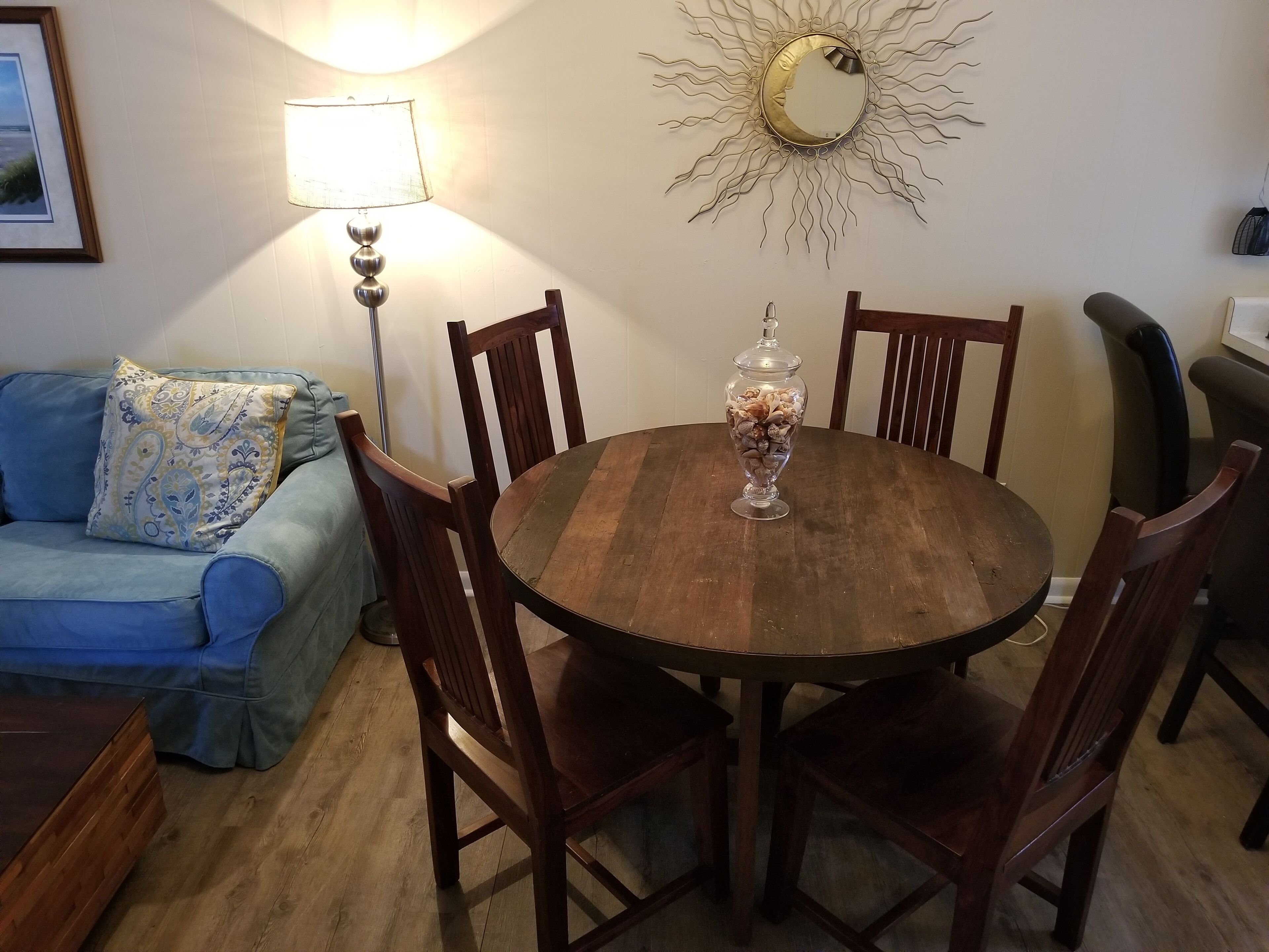 A dining area with a wooden table, four chairs, a decorative mirror, a lamp, and a blue sofa with a patterned pillow.