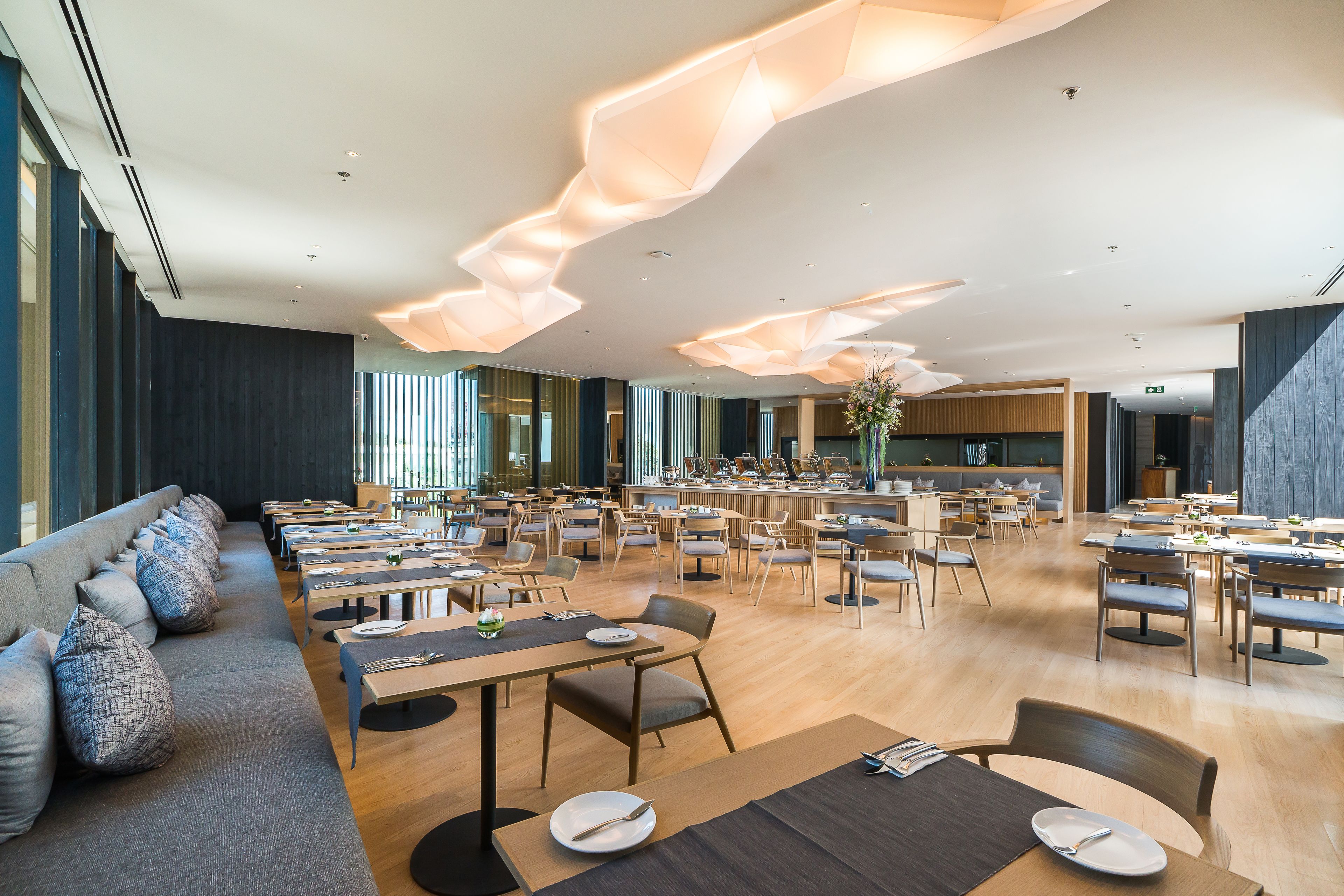A spacious dining area with multiple tables and chairs, a long bench seating area, and a modern ceiling design.