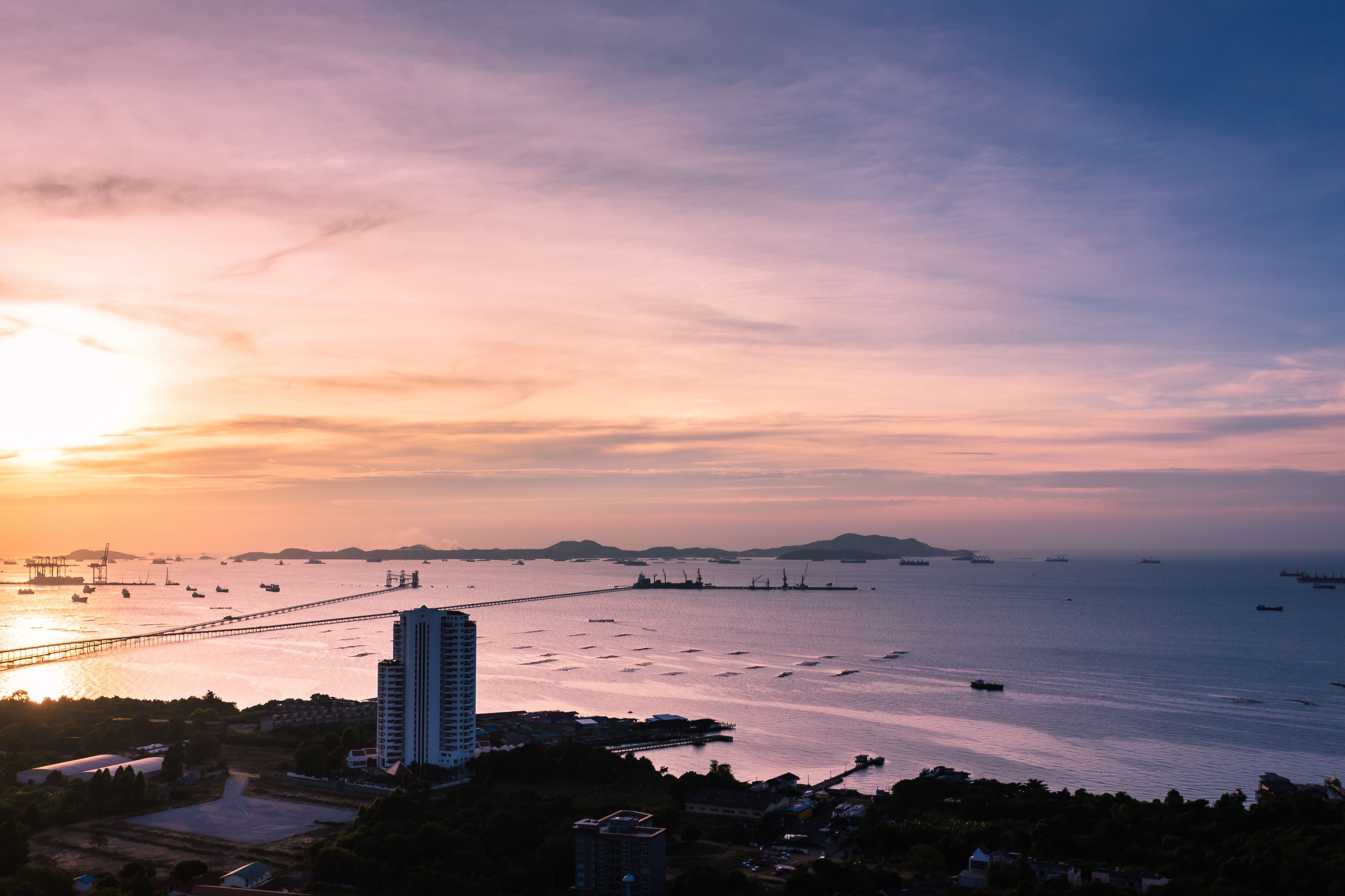 A coastal cityscape at sunset with a prominent bridge, multiple boats, and a tall building.