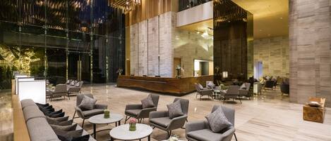 Eine geräumige Lobby mit modernem Design, inklusive einer Rezeption, gemütlichen Sitzbereichen und einer großen Glaswand mit Blick ins Innere.