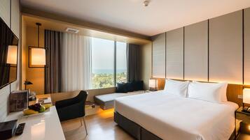 Habitación de hotel moderna con una cama grande, un escritorio con un control remoto, una silla y vistas a la ciudad a través de amplios ventanales.