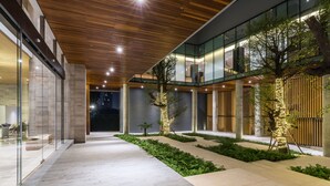 Un bâtiment moderne doté d’une façade en verre, d’un plafond en bois et d’un espace extérieur aménagé avec des allées et des arbres.