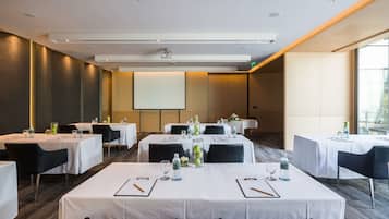 Una sala de conferencias con varias mesas, sillas y una pantalla de proyector. Las mesas están preparadas con botellas de agua, vasos y blocs de notas.