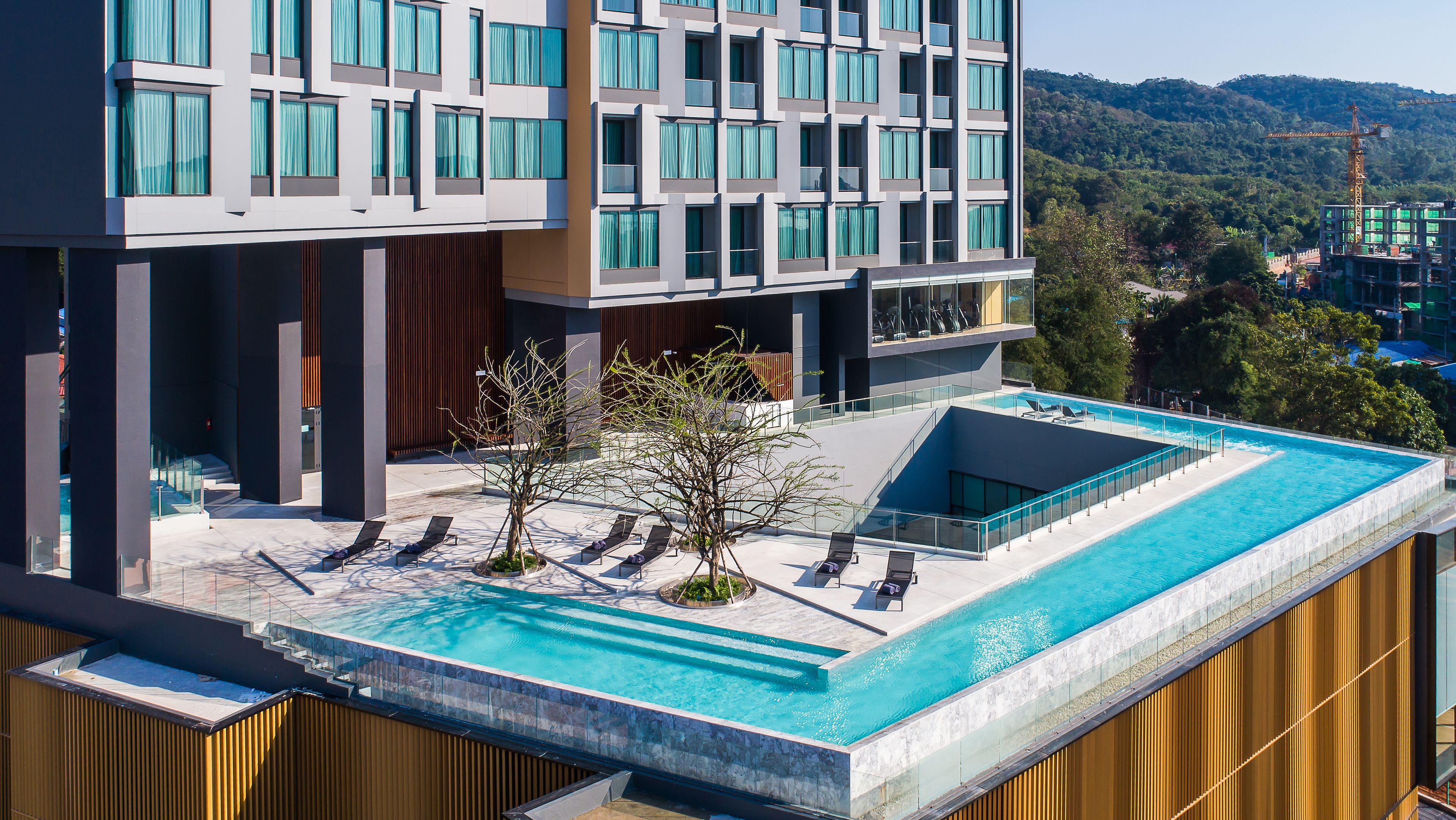 A modern hotel with a rooftop infinity pool, a landscaped terrace, and a view of a construction site in the background.