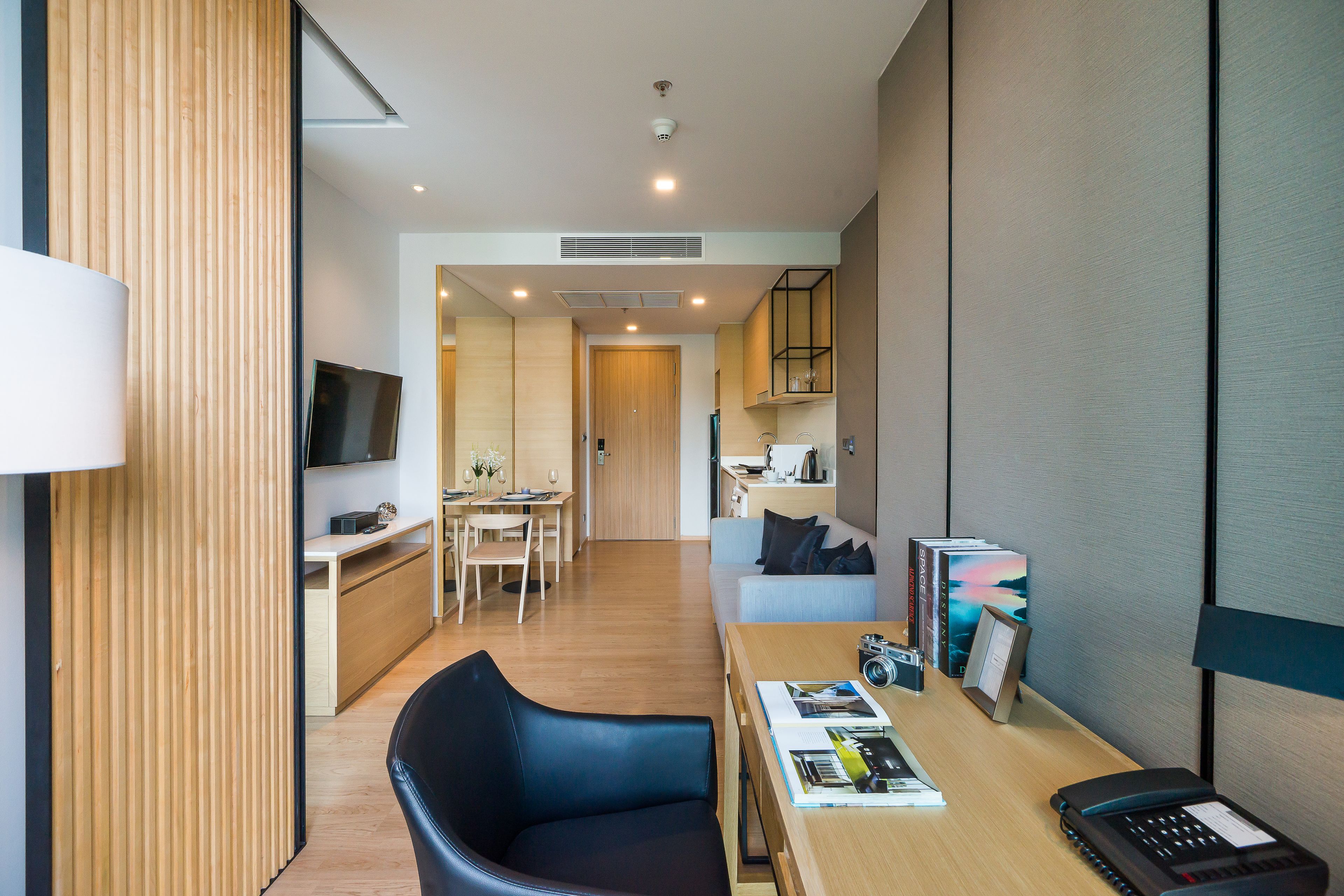 A modern hotel room with a wooden floor, a desk with a laptop, a black leather chair, a sofa, and a TV mounted on the wall.