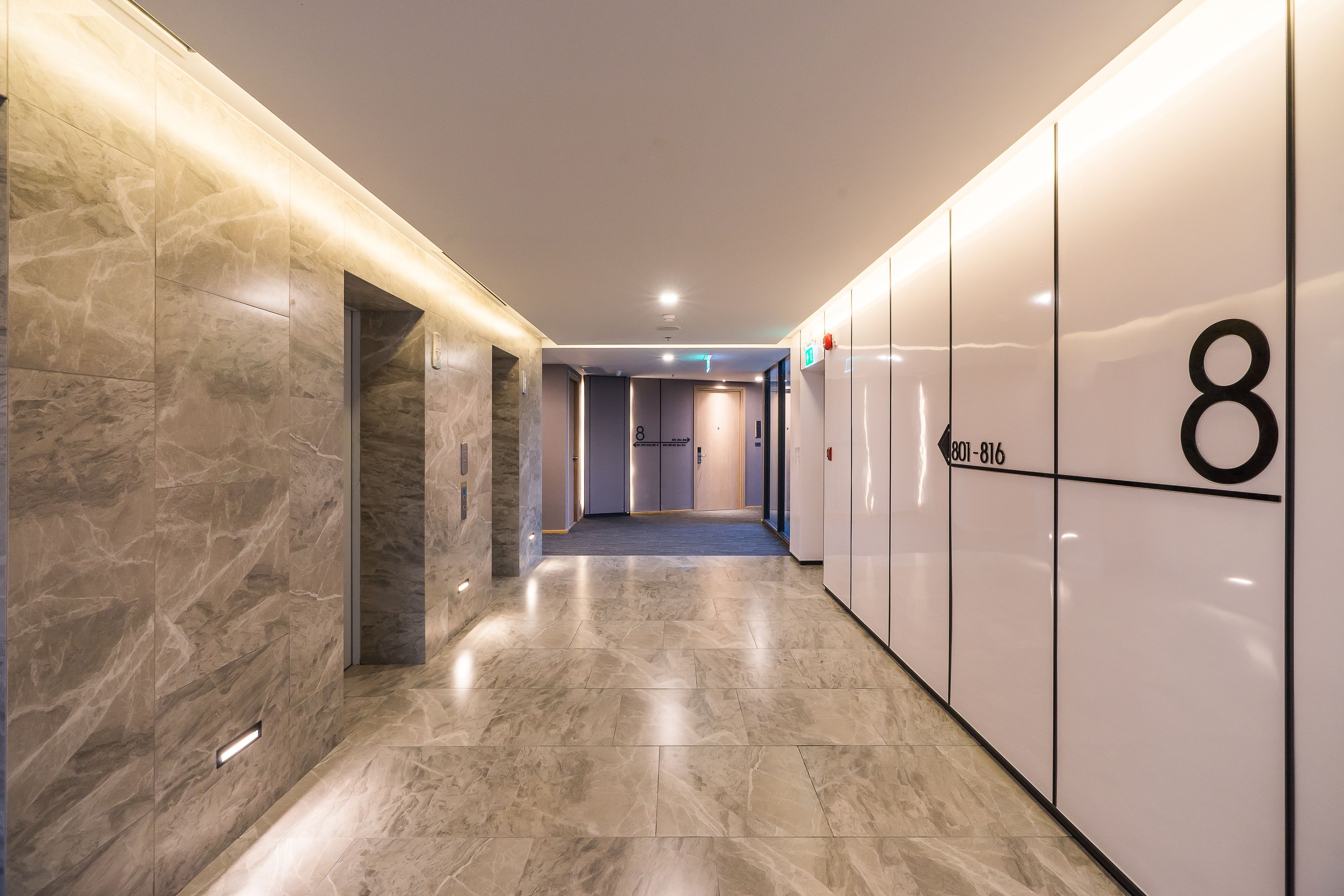 Corredor moderno com piso de mármore, placa indicando o número 8 e porta de acesso a um elevador.