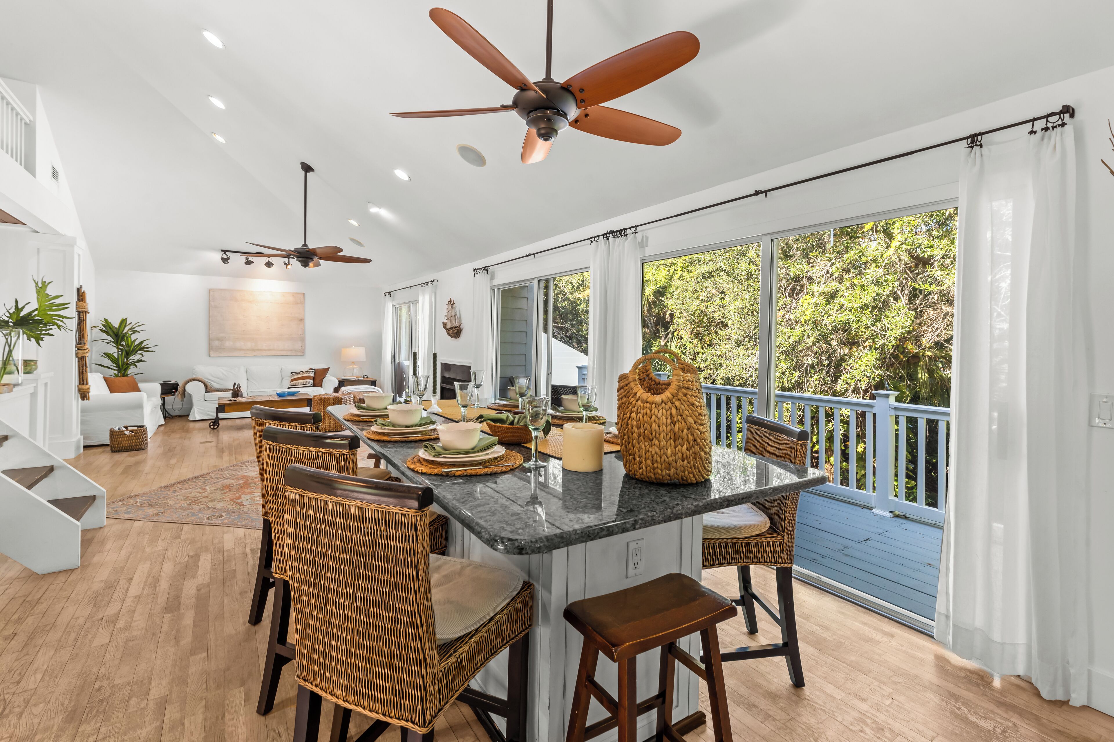 A spacious kitchen with a central island, dining area with wicker chairs, and a large window with a view of the outdoors.
