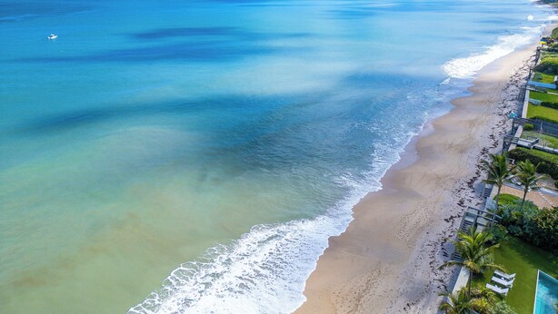 Luftaufnahme eines Strandes mit klarem Wasser, sandigem Ufer und einer Reihe von Gebäuden mit Poolbereich.