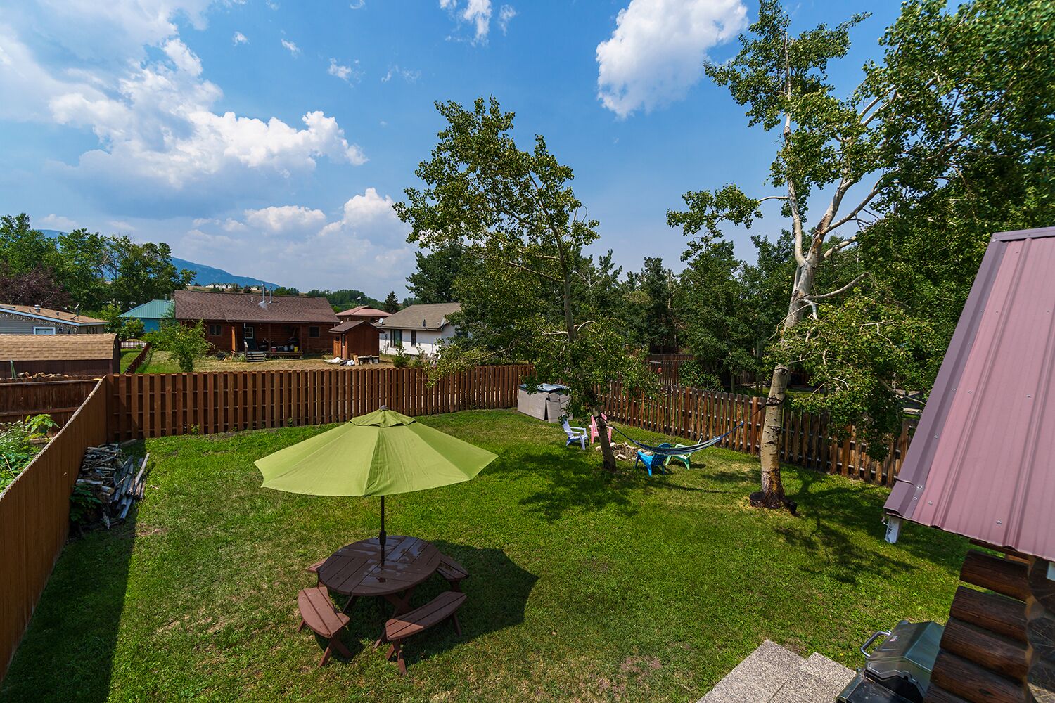 Ein Garten mit einer Holzterrasse, einem Picknicktisch mit Bänken und einem grünen Sonnenschirm.